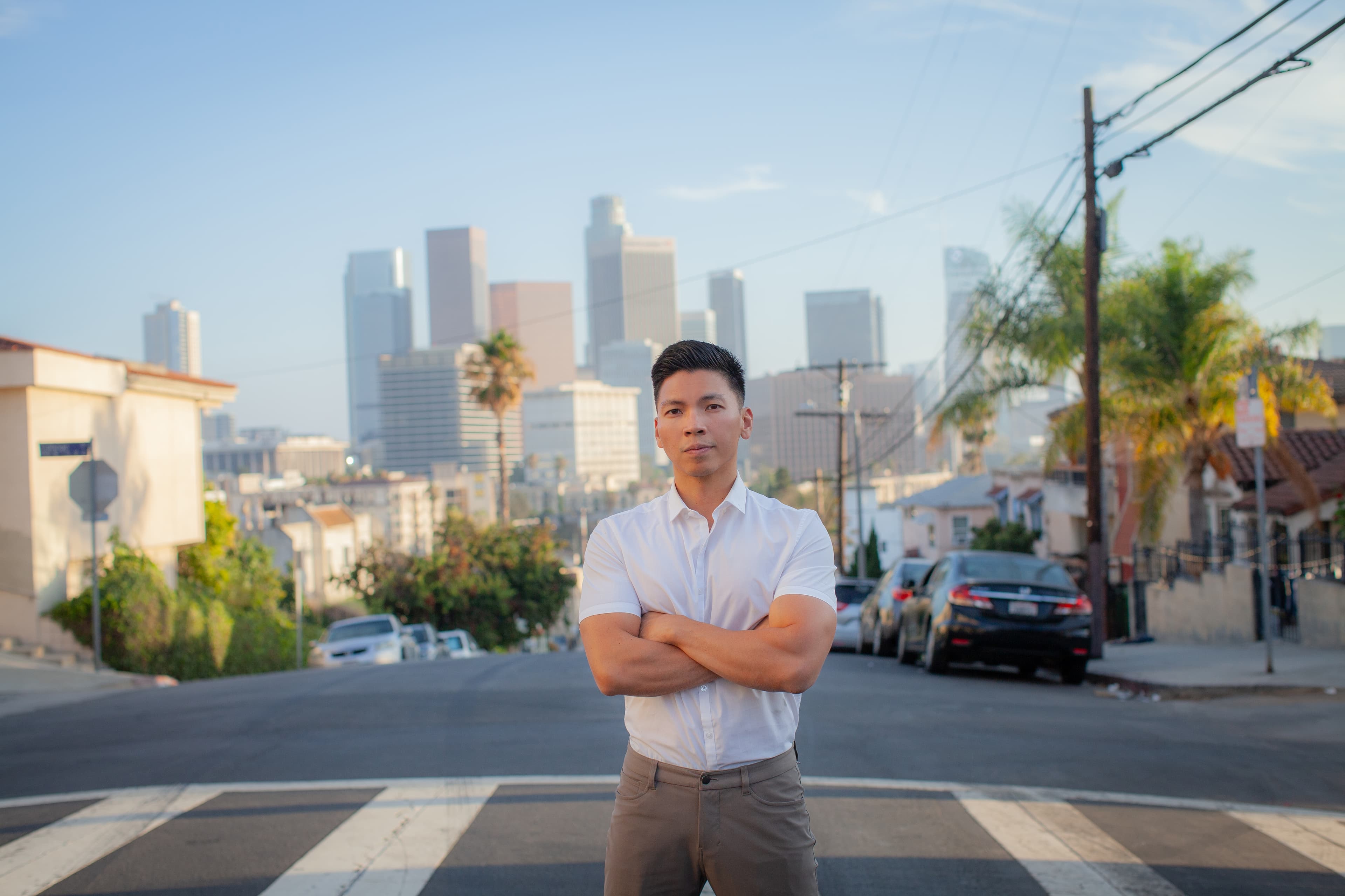 Photo of Kenneth Mejia standing in the middle of the street with the Downtown Los Angeles Skyline of Buildings in the background during the sunset. Kenneth has his arms crossed and is ready to run for re-election for LA City Controller.